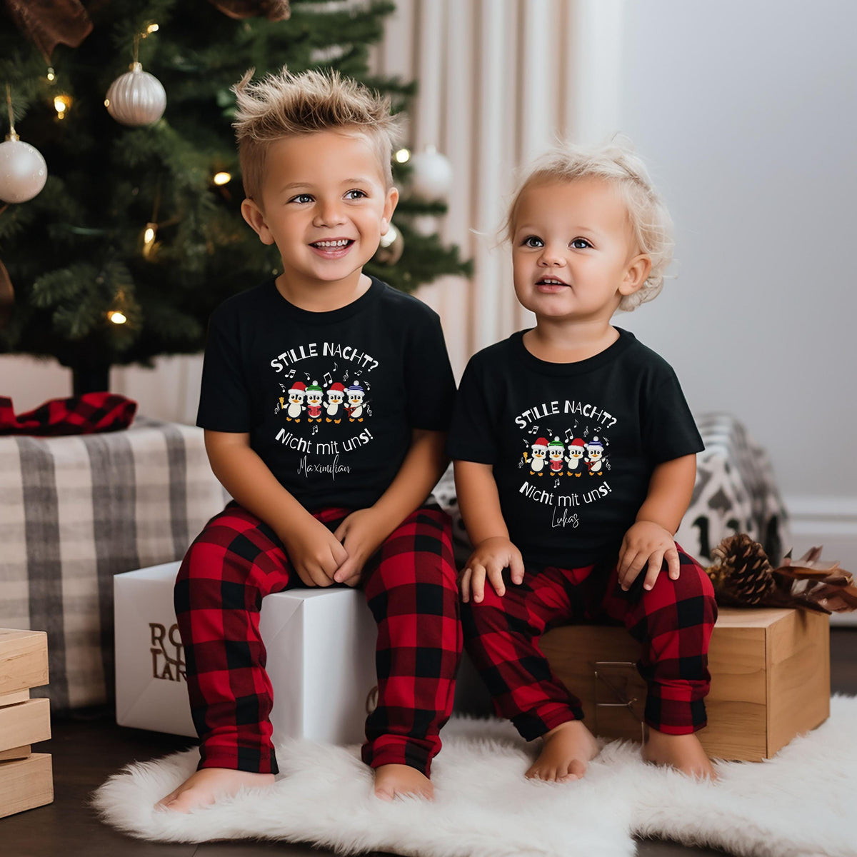 Zwei kleine Kinder sitzen barfuß auf Geschenkboxen neben einem Weihnachtsbaum und tragen schwarze kurzärmelige Oberteile im passenden Weihnachtslook. Vier Pinguine mit bunten Weihnachtsmützen und Musiknoten begleiten den Spruch „Stille Nacht? Nicht mit uns!“. Darunter sind die Beispielnamen Maximilian und Lukas gedruckt. Rot-schwarz karierte Hosen, ein weißer Fellteppich, Tannenzapfen und verpackte Geschenke ergänzen die Szene. Personalisiertes Pinguin-Outfit für Kinder und Babys mit wählbarem Namen.