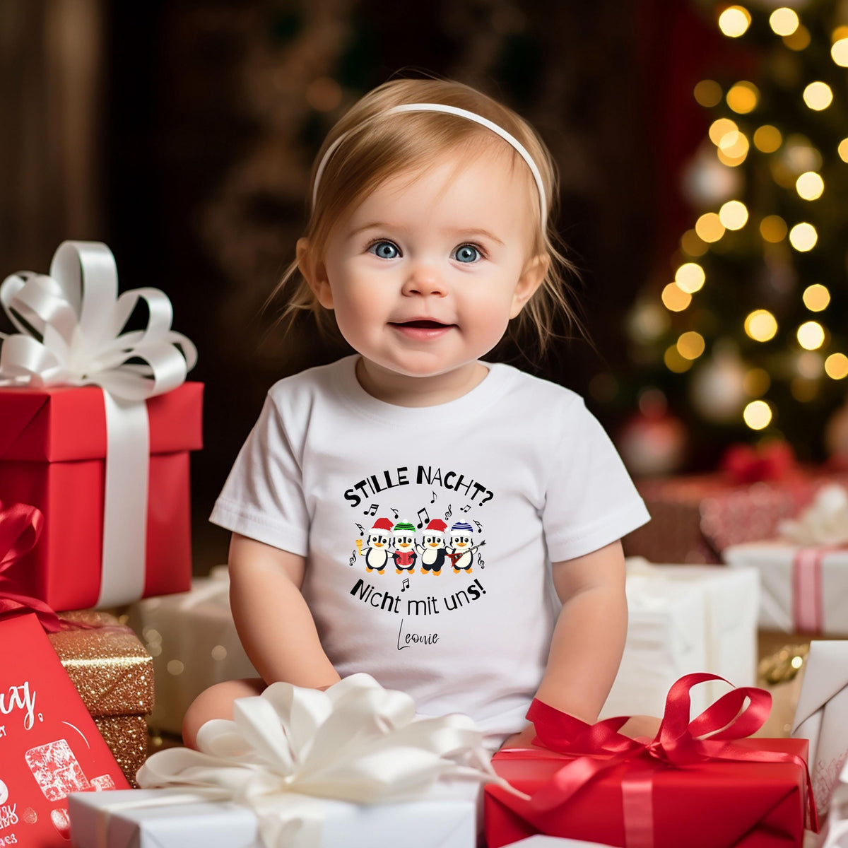 Baby im weißen kurzärmeligen Weihnachtsbody sitzt zwischen roten, weißen und goldenen Geschenkpaketen mit Schleifen. Auf der Vorderseite stehen „Stille Nacht? Nicht mit uns!“ und der Beispielname Leonie; vier Pinguine mit bunten Weihnachtsmützen und Musiknoten ergänzen den Druck. Das Baby trägt ein weißes Haarband und schaut in die Kamera. Unscharfe Lichter eines Weihnachtsbaums leuchten im Hintergrund. Personalisiertes Pinguin-Outfit für Babys, passend zu Kinder-Shirts und Pullovern im Geschwisterlook.
