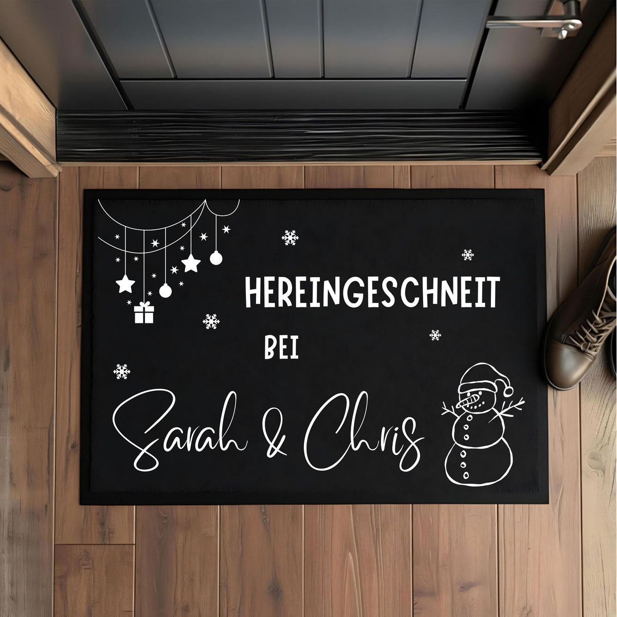 Personalisierte Weihnachtsfußmatte für Paare mit weißem Schriftzug „Hereingeschneit bei Sarah & Chris“ auf schwarzem Grund. Rechts steht ein gezeichneter Schneemann mit Weihnachtsmütze; links hängen Sterne, Kugeln und ein Geschenk, dazwischen liegen Schneeflocken. Die rechteckige Matte mit schwarzem Rand ist von oben auf einem braunen Holzboden vor einer dunklen Tür abgebildet. Ein Stiefel am rechten Bildrand ergänzt die Eingangsszene. Die Namen Sarah und Chris veranschaulichen die Wunschpersonalisierung.