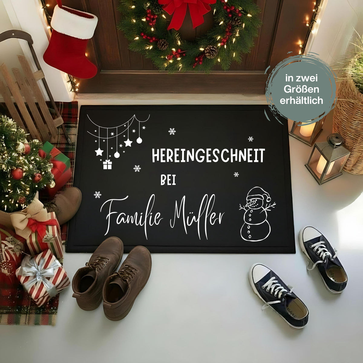 Personalisierte Weihnachtsfußmatte in Schwarz mit weißem Schriftzug „Hereingeschneit bei Familie Müller“. Schneemann mit Weihnachtsmütze, Schneeflocken und hängende Sterne, Kugeln und ein Geschenk schmücken die rechteckige Türmatte. Sie liegt vor einer Holztür mit Weihnachtskranz, neben Laternen, Schuhen, einem kleinen Tannenbaum und verpackten Geschenken. Das Bild zeigt die Namensgestaltung als Beispiel; der Hinweis „in zwei Größen erhältlich“ ergänzt die festliche Präsentation für den Eingangsbereich.