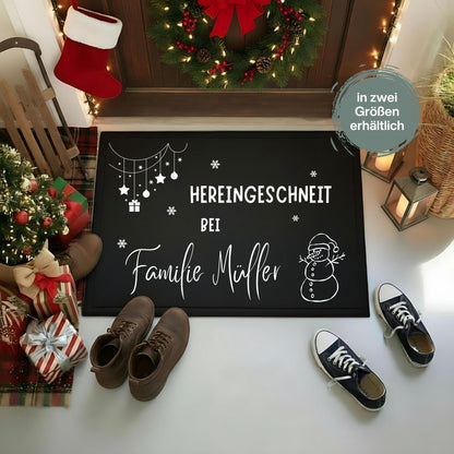 Personalisierte Weihnachtsfußmatte in Schwarz mit weißem Schriftzug „Hereingeschneit bei Familie Müller“. Schneemann mit Weihnachtsmütze, Schneeflocken und hängende Sterne, Kugeln und ein Geschenk schmücken die rechteckige Türmatte. Sie liegt vor einer Holztür mit Weihnachtskranz, neben Laternen, Schuhen, einem kleinen Tannenbaum und verpackten Geschenken. Das Bild zeigt die Namensgestaltung als Beispiel; der Hinweis „in zwei Größen erhältlich“ ergänzt die festliche Präsentation für den Eingangsbereich.