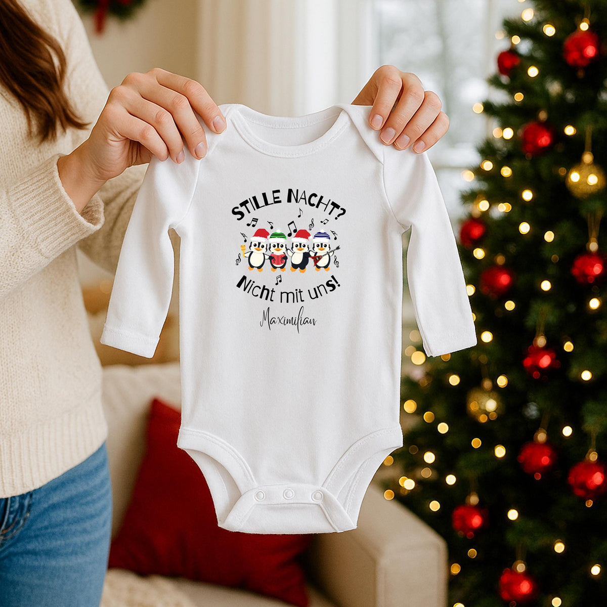 Weißer langärmeliger Babybody mit personalisiertem Pinguin-Weihnachtsmotiv, von einer Person an den Schultern hochgehalten. Vier Pinguine mit bunten Weihnachtsmützen, Musikinstrumenten und Noten stehen zwischen „Stille Nacht? Nicht mit uns!“; darunter ist der Beispielname Maximilian gedruckt. Die Vorderansicht zeigt überlappende Schultern, lange Ärmel und drei Druckknöpfe im Schritt. Im Hintergrund stehen ein rot-golden geschmückter Weihnachtsbaum und ein Sofa. Individuelles Baby-Outfit für Weihnachten.