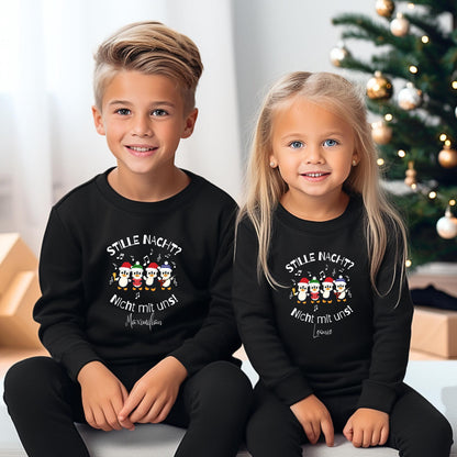 Zwei Kinder sitzen nebeneinander in schwarzen personalisierten Weihnachtspullovern mit langen Ärmeln und Rundhalsausschnitt. Das Motiv zeigt vier musikalische Pinguine mit bunten Weihnachtsmützen, Noten und dem Spruch „Stille Nacht? Nicht mit uns!“. Unter dem Druck stehen die Beispielnamen Maximilian und Leonie. Der passende Geschwisterlook wird vor einem beleuchteten Weihnachtsbaum mit Geschenken gezeigt. Hosen ergänzen die Outfits. Weihnachtskleidung für Jungen und Mädchen mit eigenem Wunschnamen.