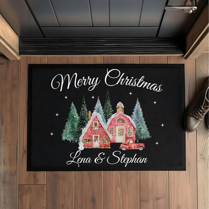 Weihnachtsfußmatte personalisiert mit „Merry Christmas“ und den Namen „Lena & Stephan“, von oben auf einem Holzboden vor einer dunklen Haustür fotografiert. Die schwarze rechteckige Matte zeigt zwei rote Häuser im Schnee, grüne und weiße Tannen, Sterne und ein rotes Auto. Ein brauner Schuh liegt rechts neben der Matte. Erhältlich in 40 × 60 oder 50 × 70 cm, mit Oberfläche aus 100 % Polypropylen und Gummirand. Individuelle Weihnachtsdeko und Geschenk für Paare, Familien oder zum Einzug in ein neues Zuhause.