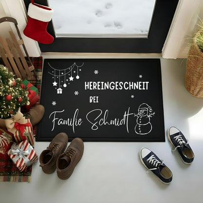 Personalisierte Weihnachtsfußmatte mit weißem Schriftzug „Hereingeschneit bei Familie Schmidt“ auf schwarzem Grund. Ein Schneemann mit Weihnachtsmütze, Schneeflocken sowie hängende Sterne, Kugeln und Geschenk ergänzen die Namensgestaltung. Die rechteckige Türmatte liegt auf hellem Boden vor einer Glastür mit Blick auf Schnee und Fußspuren. Braune Schuhe und schwarze Turnschuhe stehen davor; links sind Tannenbaum, Geschenke, Schlitten und Weihnachtsstrumpf zu sehen. Familie Schmidt ist ein Namensbeispiel.