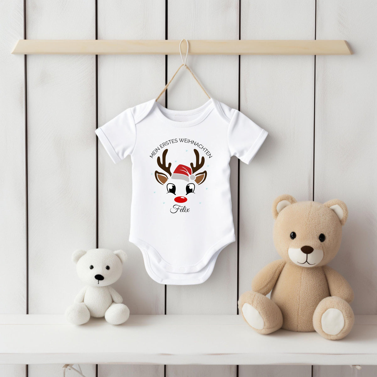 Weißer Kurzarm-Babybody mit Namen Felix hängt an einem Bügel vor einer weißen Holzpaneelwand. Der Druck zeigt ein Rentiergesicht mit braunem Geweih, roter Nase und Nikolausmütze unter dem Bogen „Mein erstes Weihnachten“. Felix steht darunter in schwarzer Schreibschrift. Auf dem weißen Regal sitzen ein weißer und ein beiger Teddybär. Die helle Produktansicht zeigt kurze Ärmel, überlappende Schultern und den Schrittverschluss des personalisierten Weihnachtsbodys aus 100 % Bio-Baumwolle für Babys erstes Fest.