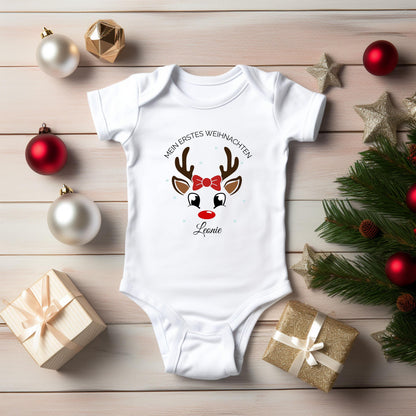 Weißer Kurzarm-Babybody mit Namen Leonie liegt auf einer hellen Holzfläche zwischen roten und weißen Christbaumkugeln, goldenen Sternen und Geschenken. Der Weihnachtsdruck zeigt ein Rentiergesicht mit Geweih, großen Augen, roter Nase und Schleife unter „Mein erstes Weihnachten“. Leonie steht darunter in schwarzer Schreibschrift. Tannenzweige mit Zapfen rahmen die Produktansicht. Kurze Ärmel und überlappende Schultern sind sichtbar. Personalisiertes Babyoutfit aus 100 % Bio-Baumwolle für das erste Fest.