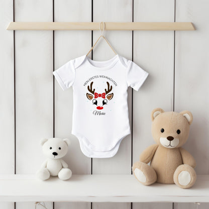 Weißer Kurzarm-Babybody mit Namen Marie hängt an einem Bügel vor einer weißen Holzpaneelwand. Der Druck zeigt ein Rentiergesicht mit braunem Geweih, roter Nase und roter Schleife unter dem Bogen „Mein erstes Weihnachten“. Marie steht darunter in schwarzer Schreibschrift. Auf dem weißen Regal sitzen ein weißer und ein beiger Teddybär. Die helle Produktansicht zeigt kurze Ärmel, überlappende Schultern und den Schrittverschluss des personalisierten Weihnachtsbodys aus 100 % Bio-Baumwolle für Babys erstes Fest.
