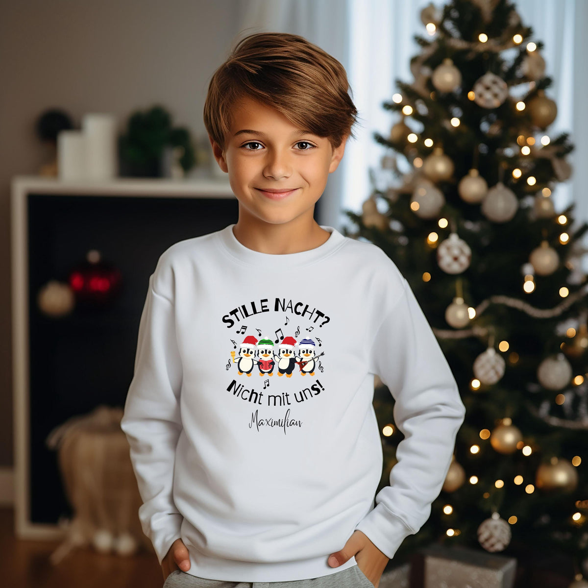 Junge im weißen personalisierten Weihnachtspullover vor einem geschmückten Weihnachtsbaum. Auf dem Sweatshirt stehen „Stille Nacht? Nicht mit uns!“ und der Beispielname Maximilian. Vier Pinguine mit bunten Weihnachtsmützen und Musikinstrumenten bilden das fröhliche Druckmotiv, ergänzt durch Noten. Vorderansicht des Kinder-Outfits mit Rundhalsausschnitt und langen Ärmeln, kombiniert mit einer grauen Hose. Geschenkidee für Kinder und ein Geschwisterlook zu Weihnachten; Name individuell wählbar. Im Wohnzimmer.