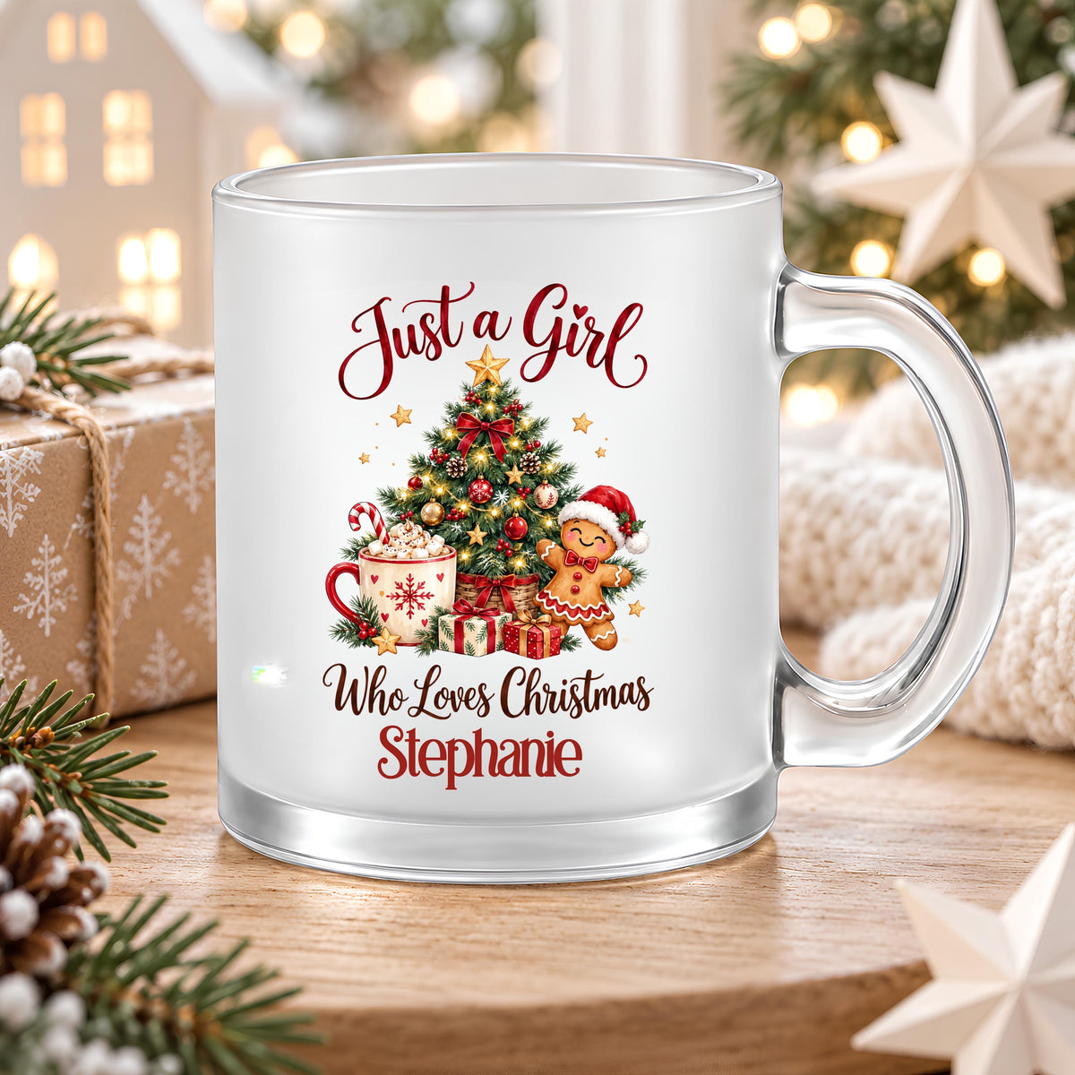 Personalisierte Weihnachtstasse mit dem Beispielnamen Stephanie auf einem Holztisch neben einem Geschenk mit Schneeflockenpapier. Auf satiniertem Glas steht „Just a Girl Who Loves Christmas“ mit geschmücktem Tannenbaum, Kakao, Zuckerstange, Lebkuchenmann und Geschenken. Der klare Henkel und Glasboden sind sichtbar; Tannenzweige, Zapfen, Strickdecke und weiße Sterne rahmen die Tasse ein. Die bedruckte 300-ml-Glastasse mit Wunschname eignet sich als Weihnachtsgeschenk für Freundin, Schwester oder Kollegin.