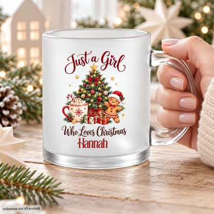 Nahaufnahme einer Hand am transparenten Henkel der personalisierten Weihnachtstasse mit dem Beispielnamen Hannah. Der rote Schriftzug „Just a Girl Who Loves Christmas“ umrahmt Tannenbaum, Kakao mit Zuckerstange, Lebkuchenmann und Geschenke auf satiniertem Glas. Der Glasboden und die Frontansicht des Drucks sind gut sichtbar. Die 300-ml-Tasse steht auf einem Holztisch zwischen Tannenzweigen, Zapfen und Sternen. Ein Weihnachtsbecher mit Wunschname als Geschenk für Freundin, Schwester oder Kollegin im Advent.