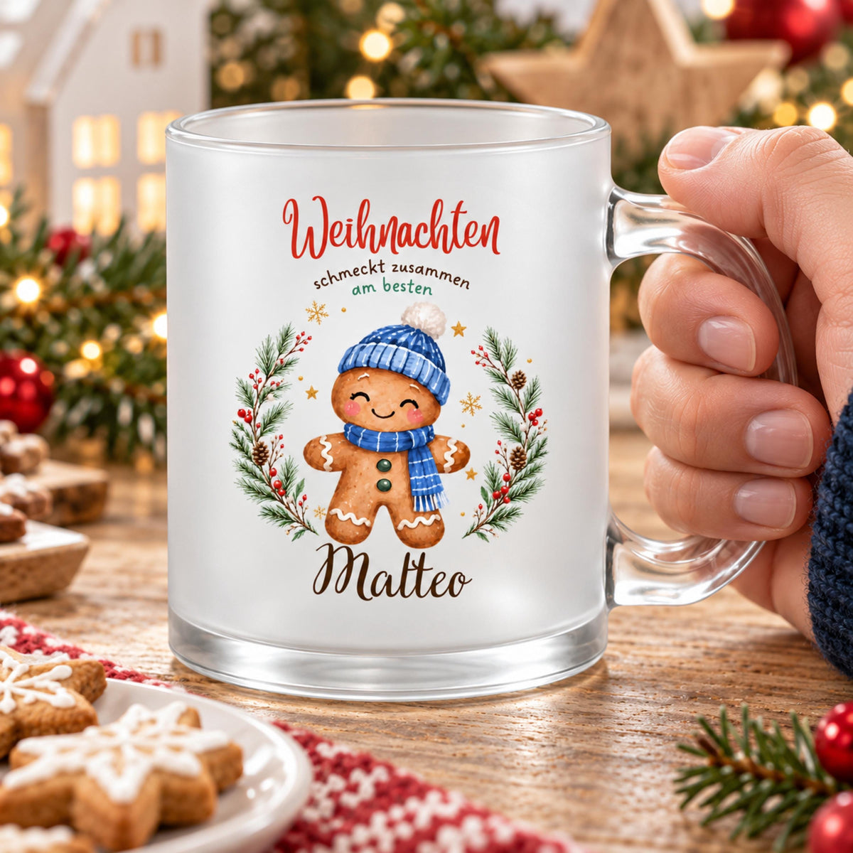 Nahaufnahme einer Hand am transparenten Henkel der Weihnachtstasse mit Lebkuchenfigur, blauer Mütze und Schal. Im Hintergrund ist Weihnachtsdekoration zu sehen. Die Lebkuchenmotive sind von einem Tannenkranz mit Zapfen, Beeren und goldenen Sternen umrahmt und tragen „Weihnachten schmeckt zusammen am besten“. Die satinierte 300-ml-Glastasse wird beidseitig mit Wunschname bedruckt. Persönliches Weihnachtsgeschenk für Familie, Freunde oder Kollegen, auch zu Nikolaus oder zum Wichteln. Im Advent. Für Kaffee.