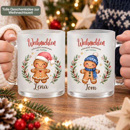 Zwei personalisierte Weihnachtstassen werden nebeneinander in Händen gehalten. Die Nahaufnahme zeigt die Lebkuchenfiguren, Wunschbeschriftungen und transparenten Glasgriffe. Die Lebkuchenmotive sind von einem Tannenkranz mit Zapfen, Beeren und goldenen Sternen umrahmt und tragen „Weihnachten schmeckt zusammen am besten“. Die satinierte 300-ml-Glastasse wird beidseitig mit Wunschname bedruckt. Persönliches Weihnachtsgeschenk für Familie, Freunde oder Kollegen, auch zu Nikolaus oder zum Wichteln. Im Advent.