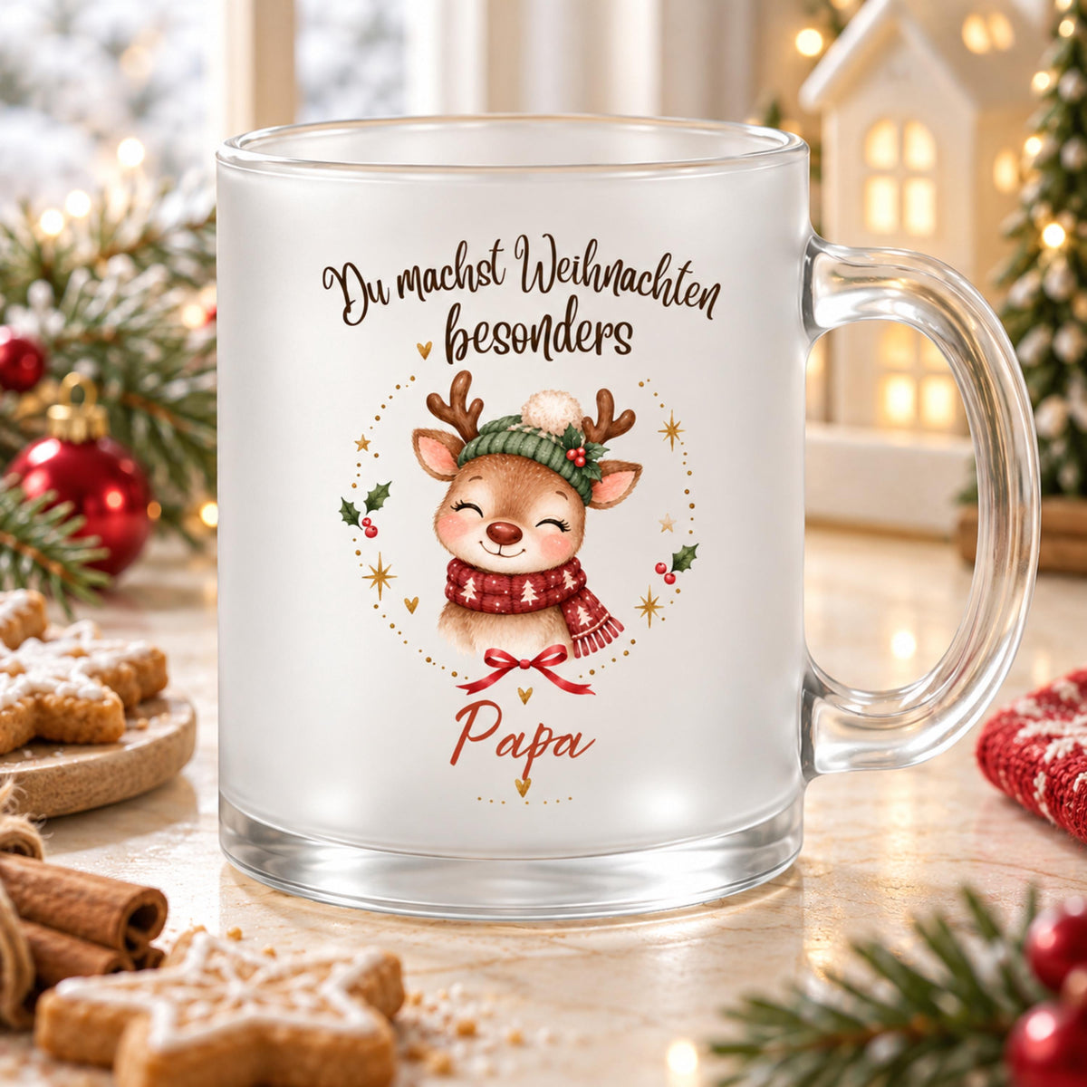 Personalisierte Rentier-Weihnachtstasse für Papa aus satiniertem Glas mit transparentem Henkel rechts. Das Rentier trägt eine grüne Strickmütze mit weißem Bommel und einen roten Schal mit weißen Tannen. Goldene Sterne, Herzen, rote Beeren und eine Schleife umrahmen das Motiv. Darüber steht „Du machst Weihnachten besonders“, darunter Papa in roter Schrift. Die Glastasse steht auf hellem Holz zwischen Sternplätzchen, Zimtstangen, Tannengrün und roten Kugeln vor einem Dekohaus. Geschenkidee für den Vater.