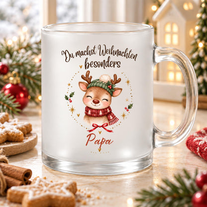 Personalisierte Rentier-Weihnachtstasse für Papa aus satiniertem Glas mit transparentem Henkel rechts. Das Rentier trägt eine grüne Strickmütze mit weißem Bommel und einen roten Schal mit weißen Tannen. Goldene Sterne, Herzen, rote Beeren und eine Schleife umrahmen das Motiv. Darüber steht „Du machst Weihnachten besonders“, darunter Papa in roter Schrift. Die Glastasse steht auf hellem Holz zwischen Sternplätzchen, Zimtstangen, Tannengrün und roten Kugeln vor einem Dekohaus. Geschenkidee für den Vater.