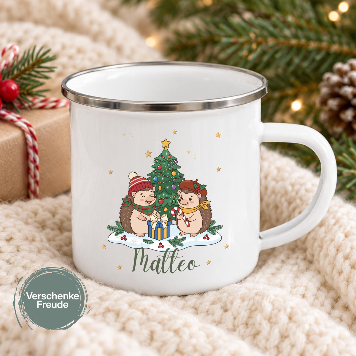Personalisierte Weihnachtstasse für Kinder mit zwei Igeln in Winterkleidung vor einem Weihnachtsbaum, Geschenk und Zuckerstange. Unter dem Motiv steht der Beispielname Matteo. Weiße Emaille-Tasse mit silberfarbenem Rand und weißem C-Henkel, ca. 300 ml. Kinderbecher mit Wunschname als Weihnachtsgeschenk oder Nikolausgeschenk für Kinder, Enkelkinder und Patenkinder. Auf einer hellen Decke neben Geschenk, Tannenzweigen und Lichtern gezeigt. Geliefert wird eine Tasse mit gewähltem Motiv und Namen.
