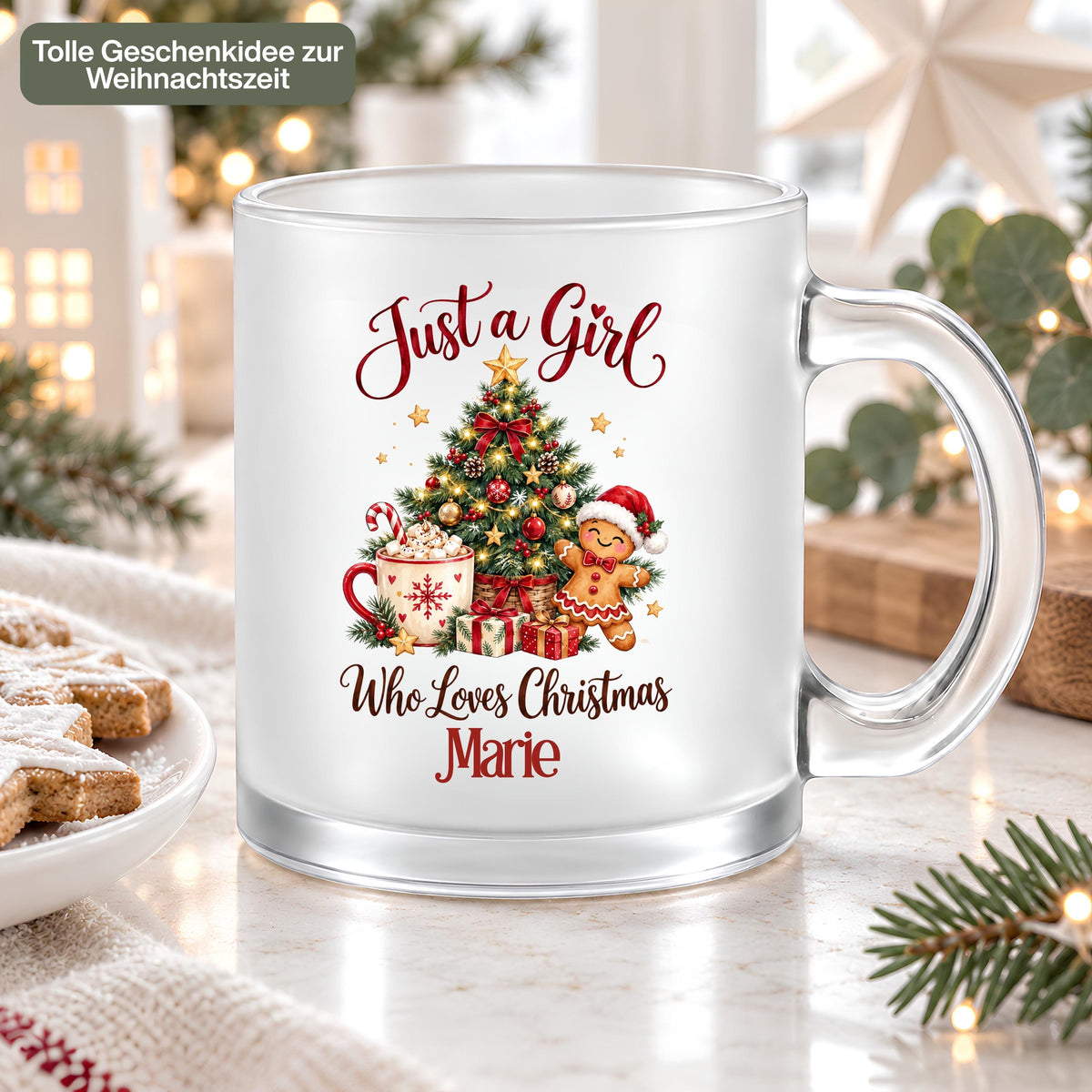 Personalisierte Weihnachtstasse aus satiniertem Glas mit dem Beispielnamen Marie auf einem Tisch neben Plätzchen und einer Strickdecke. Der Aufdruck „Just a Girl Who Loves Christmas“ zeigt Tannenbaum, Kakao, Zuckerstange, Lebkuchenmann und Geschenke. Transparenter Henkel und Glasboden sind sichtbar; Sterne, Tannenzweige und Lichter schmücken die Umgebung. Oben steht „Tolle Geschenkidee zur Weihnachtszeit“. Die 300-ml-Glastasse mit Wunschname ist ein Weihnachtsgeschenk für Freundin, Schwester oder Kollegin.
