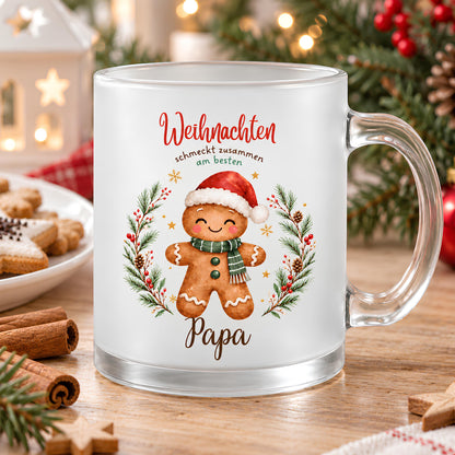 Personalisierte Weihnachtstasse mit einer Lebkuchenfigur mit Zipfelmütze und Schal, präsentiert in weihnachtlicher Umgebung mit Tannenzweigen und Lichtern. Die Lebkuchenmotive sind von einem Tannenkranz mit Zapfen, Beeren und goldenen Sternen umrahmt und tragen „Weihnachten schmeckt zusammen am besten“. Die satinierte 300-ml-Glastasse wird beidseitig mit Wunschname bedruckt. Persönliches Weihnachtsgeschenk für Familie, Freunde oder Kollegen, auch zu Nikolaus oder zum Wichteln. Für Kaffee, Tee und Kakao.