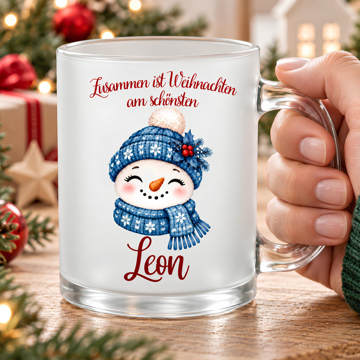 Nahaufnahme einer personalisierten Weihnachtstasse aus satiniertem Glas mit dem Namen Leon in roter Schreibschrift. Eine Hand hält den transparenten Henkel rechts. Der lächelnde Schneemann trägt eine blaue Strickmütze mit weißem Bommel und Schneeflocken sowie einen passenden blauen Schal. Darüber steht „Zusammen ist Weihnachten am schönsten“. Die Glastasse steht auf einem Holztisch; rote Weihnachtskugeln, Tannengrün, Geschenke und warme Lichter bilden den unscharfen Hintergrund. Höhe ca. 95 mm.