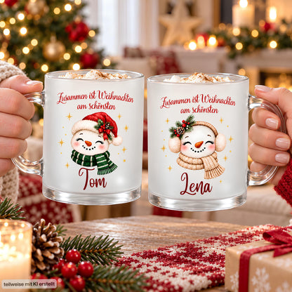 Zwei Hände halten personalisierte Schneemann-Weihnachtstassen mit Namen Tom und Lena an den transparenten Glashenkeln. Tom zeigt Nikolausmütze und grünen Schal, Lena beige Ohrenschützer und Schal. Über beiden Motiven steht „Zusammen ist Weihnachten am schönsten“. Die satinierten Glastassen sind mit einem Getränk und Sahnehaube gefüllt. Vor dem Weihnachtsbaum liegen Tannengrün, Zapfen, rote Beeren, eine Kerze, ein Geschenk und rot-weißer Strickstoff auf dem Tisch. Geschenkidee für Paare im Advent.