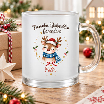 Personalisierte Rentier-Weihnachtstasse mit Namen Felix aus satiniertem Glas und transparentem Henkel rechts. Das Rentier trägt eine Nikolausmütze und einen blauen Schal mit weißen Tannen. Goldene Sterne, Herzen, eine rote Schleife und Beerenzweige umrahmen das Motiv unter „Du machst Weihnachten besonders“. Felix steht darunter in roter Schrift. Auf dem Holztisch liegen Tannengrün, Christbaumkugeln und ein Holzstern neben einem Geschenk. Lichter und ein Dekohaus begleiten die Weihnachtsgeschenkidee.
