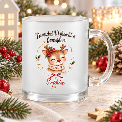 Personalisierte Rentier-Weihnachtstasse mit Namen Sophia aus satiniertem Glas, mit transparentem Henkel rechts. Das Rentier trägt einen beigen Schal mit roten Herzen und eine rote Schleife am Geweih. Goldene Sterne, Herzen, Tannengrün und rote Beeren umrahmen das Motiv. Darüber steht „Du machst Weihnachten besonders“, darunter Sophia in roter Schrift. Die Glastasse steht auf Holz zwischen Christbaumkugeln, Zapfen und Strickstoff. Ein Dekohaus und eine Kerze schaffen Weihnachtsstimmung als Geschenkidee.