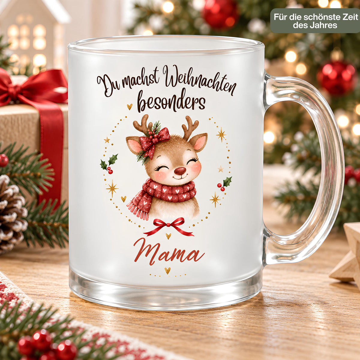 Weihnachtstasse für Mama aus satiniertem Glas mit Rentier und transparentem Henkel rechts. Das Rentier trägt einen roten Herzschal und eine rote Schleife am Geweih, mit Tannengrün. Goldene Sterne, Herzen und Beerenzweige umrahmen das Motiv unter „Du machst Weihnachten besonders“. Mama steht darunter in Rot. Auf dem Holztisch begleiten Geschenke mit rotem Band, Zapfen, Tannenzweige und ein Holzstern die Tasse; im Hintergrund leuchtet der Baum. Ein Schild betont „Für die schönste Zeit des Jahres“.