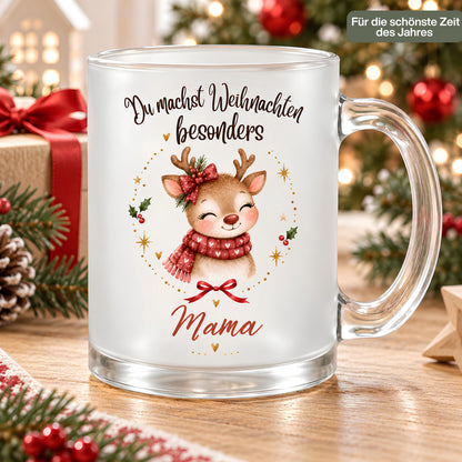 Weihnachtstasse für Mama aus satiniertem Glas mit Rentier und transparentem Henkel rechts. Das Rentier trägt einen roten Herzschal und eine rote Schleife am Geweih, mit Tannengrün. Goldene Sterne, Herzen und Beerenzweige umrahmen das Motiv unter „Du machst Weihnachten besonders“. Mama steht darunter in Rot. Auf dem Holztisch begleiten Geschenke mit rotem Band, Zapfen, Tannenzweige und ein Holzstern die Tasse; im Hintergrund leuchtet der Baum. Ein Schild betont „Für die schönste Zeit des Jahres“.