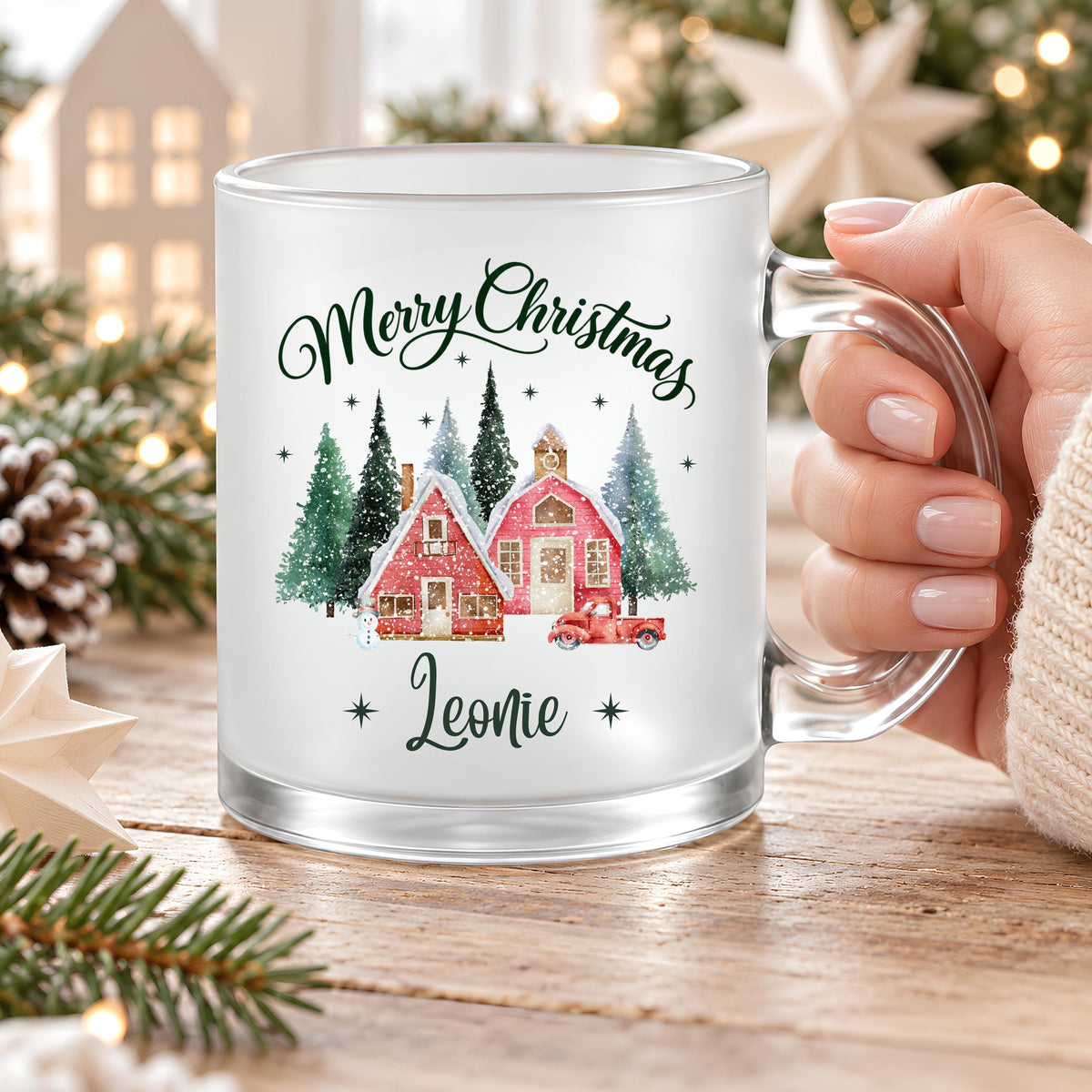 Eine Hand hält die personalisierte Weihnachtstasse mit Namen Leonie am transparenten Glashenkel. Der satinierte Weihnachtsbecher trägt Merry Christmas und ein rotes Weihnachtsdorf mit Tannen. Tasse mit Wunschname und ca. 300 ml Fassungsvermögen für Kaffee, Tee, Kakao oder Glühwein im Advent. Eine persönliche Weihnachtsgeschenkidee für die beste Freundin, Mama, Oma oder Kollegin, geeignet als Nikolausgeschenk oder Wichtelgeschenk. Individuelle Glastasse für Genussmomente zur Weihnachtszeit. Höhe ca. 95 mm.