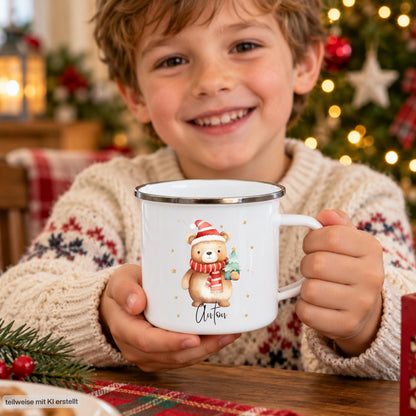 Personalisierte Weihnachtstasse für Kinder aus weißer Emaille mit silberfarbenem Rand und weißem Henkel. Das Motiv zeigt einen braunen Weihnachtsbären mit roter Weihnachtsmütze, rot-weiß gestreiftem Schal und einem grünen Tannenbaum im Arm. Unter dem Bären steht ein Beispielname; die Kindertasse wird mit Wunschname bedruckt. Weihnachtsbecher für Bärenfans und eine Geschenkidee zu Weihnachten oder Nikolaus für Kinder, Enkelkinder und Patenkinder. Ein Junge zeigt die Tasse mit Namensdruck.