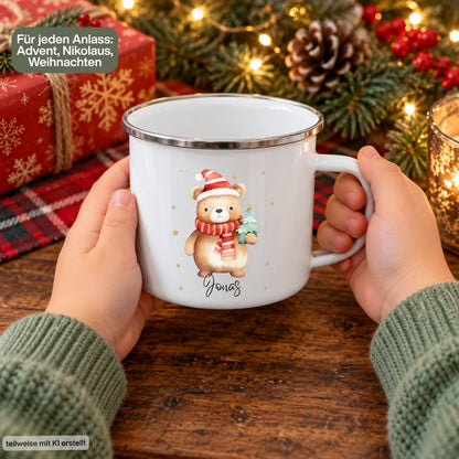 Personalisierte Weihnachtstasse für Kinder aus weißer Emaille mit silberfarbenem Rand und weißem Henkel. Das Motiv zeigt einen braunen Weihnachtsbären mit roter Weihnachtsmütze, rot-weiß gestreiftem Schal und einem grünen Tannenbaum im Arm. Unter dem Bären steht ein Beispielname; die Kindertasse wird mit Wunschname bedruckt. Weihnachtsbecher für Bärenfans und eine Geschenkidee zu Weihnachten oder Nikolaus für Kinder, Enkelkinder und Patenkinder. Ansicht der Tasse zwischen zwei Händen.