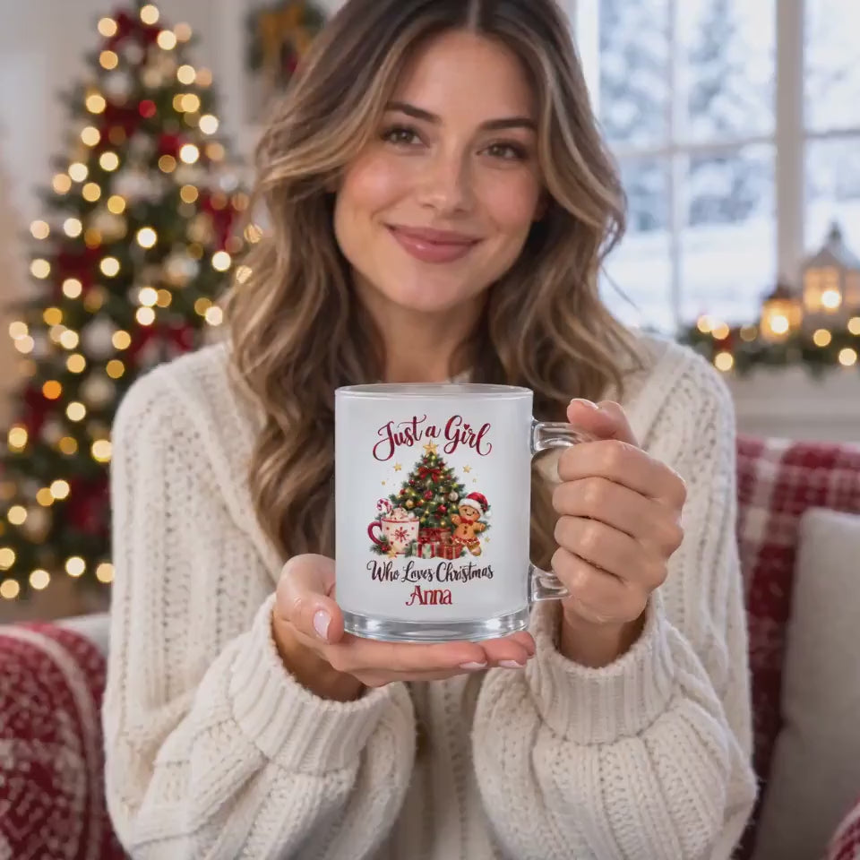 Eine Frau präsentiert die personalisierte Weihnachtstasse aus satiniertem Glas mit dem Beispielnamen Anna vor einem beleuchteten Weihnachtsbaum. Auf der 300-ml-Glastasse steht „Just a Girl Who Loves Christmas“ über und unter einem festlichen Motiv mit Tannenbaum, Kakao, Zuckerstange, Lebkuchenmann und Geschenken. Das Video zeigt den transparenten Henkel und die Ansicht des Aufdrucks. Ein Weihnachtsbecher mit Wunschname als persönliches Weihnachtsgeschenk für Freundin, Schwester oder Kollegin im Advent.